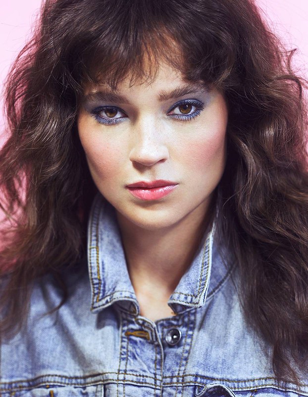 Close-up portrait of a woman with voluminous brown curly hair wearing a denim jacket against a pink background