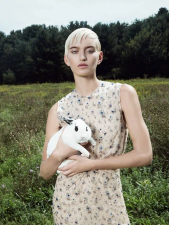 Woman with short blonde hair holds a white rabbit with black spots in a field with trees under an overcast sky in an eye-level shot.