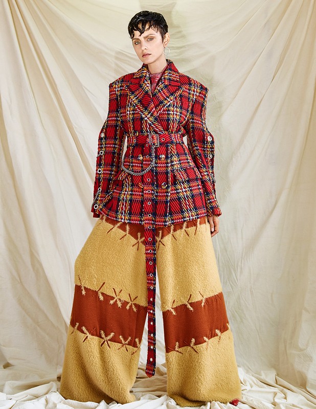 A model in a red and gold plaid jacket with gold and brown patchwork pants poses against an off-white backdrop in a studio setting.