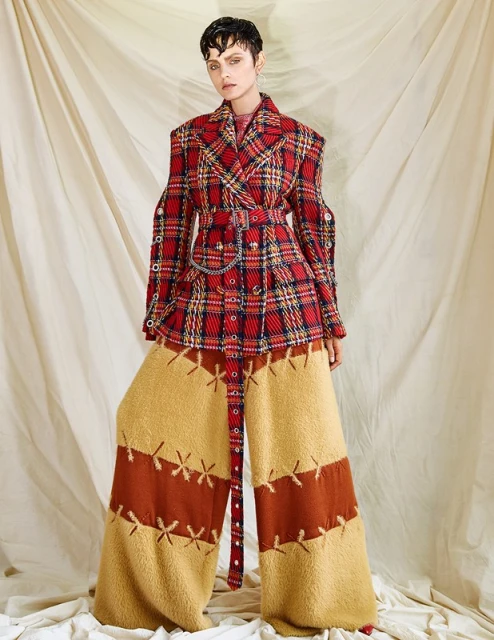 A model in a red and gold plaid jacket with gold and brown patchwork pants poses against an off-white backdrop in a studio setting.