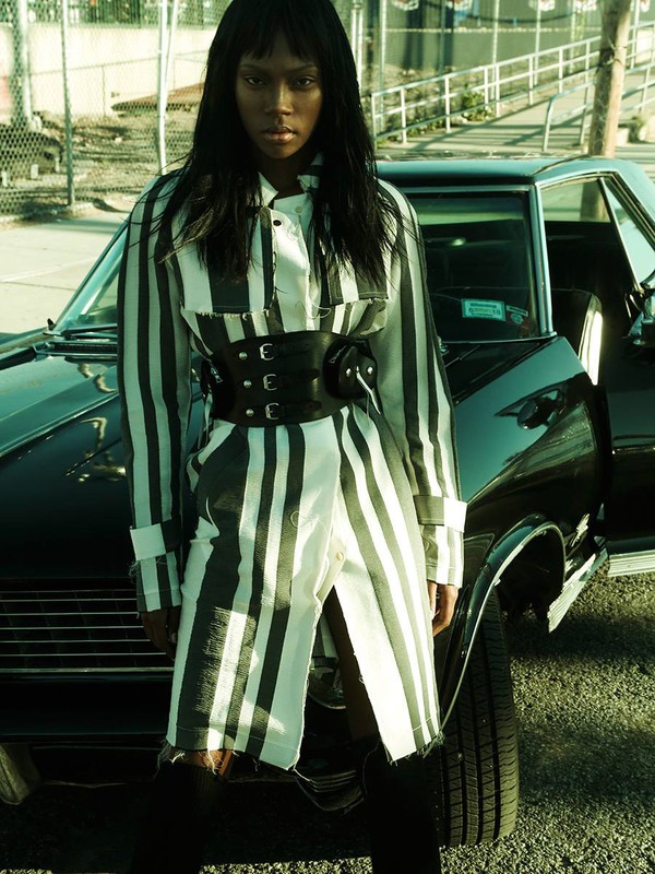 A woman in a striped jacket and corset poses next to a black car on an outdoor city street in daytime for a fashion editorial.