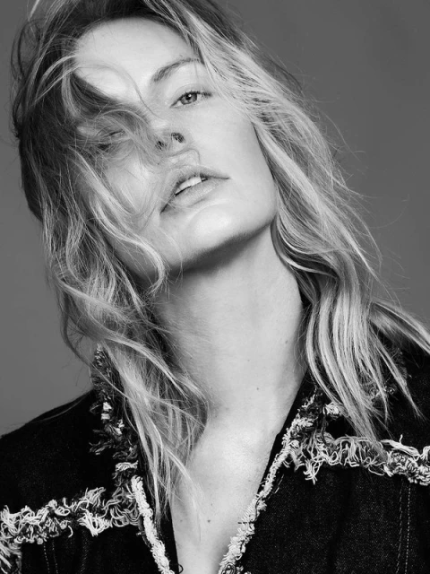 Black and white portrait of a woman with light, wavy hair wearing a fringed denim jacket against a neutral background in a studio.