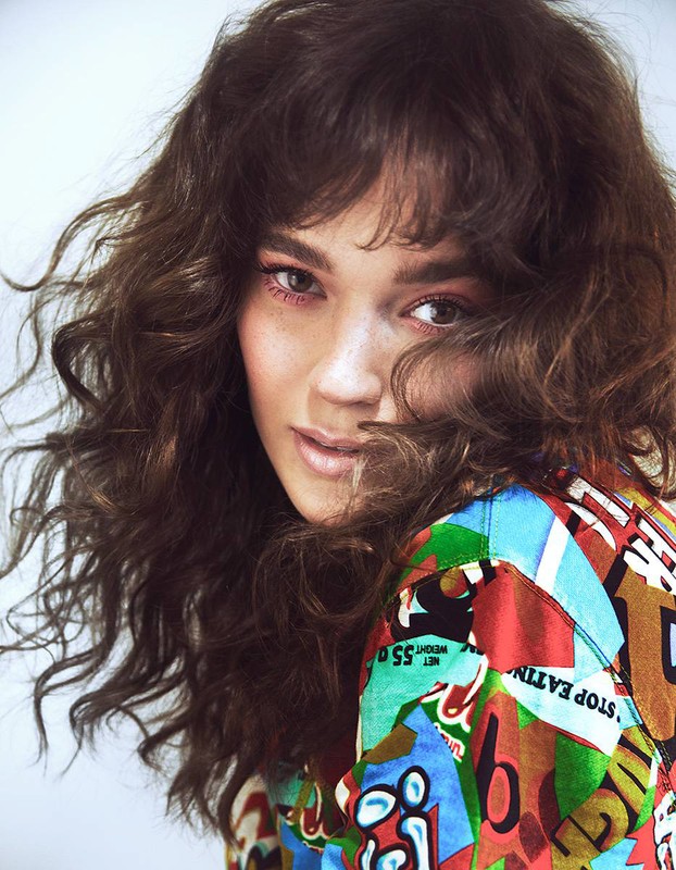 Close-up portrait of a young woman with wavy brown hair, wearing a colorful patterned shirt, looking at the camera with a slight smile.