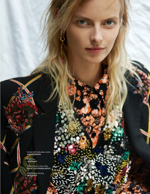 A stylish adult female poses in a studio wearing a black floral embroidered jacket and floral print shirt with sequin embellishments.