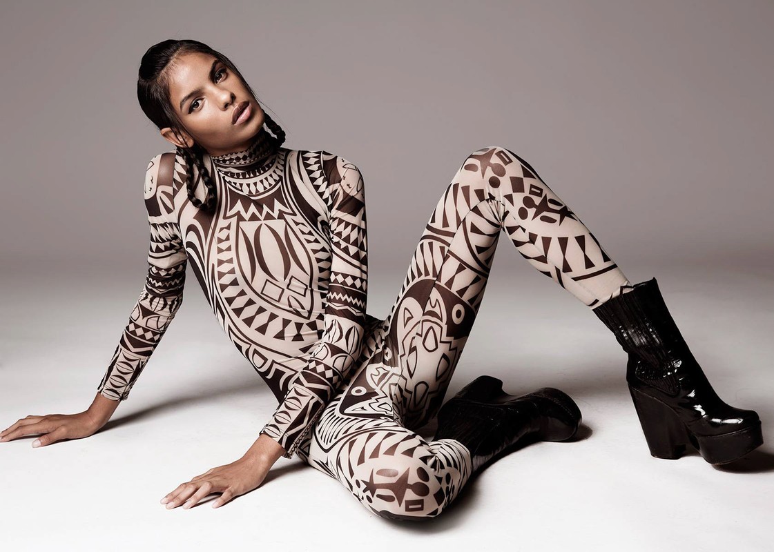 A woman models a patterned bodysuit and black platform boots, posing on the floor in a fashion forward studio portrait.