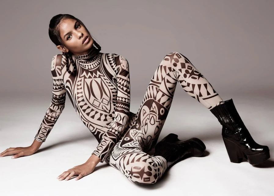 A woman models a patterned bodysuit and black platform boots, posing on the floor in a fashion forward studio portrait.