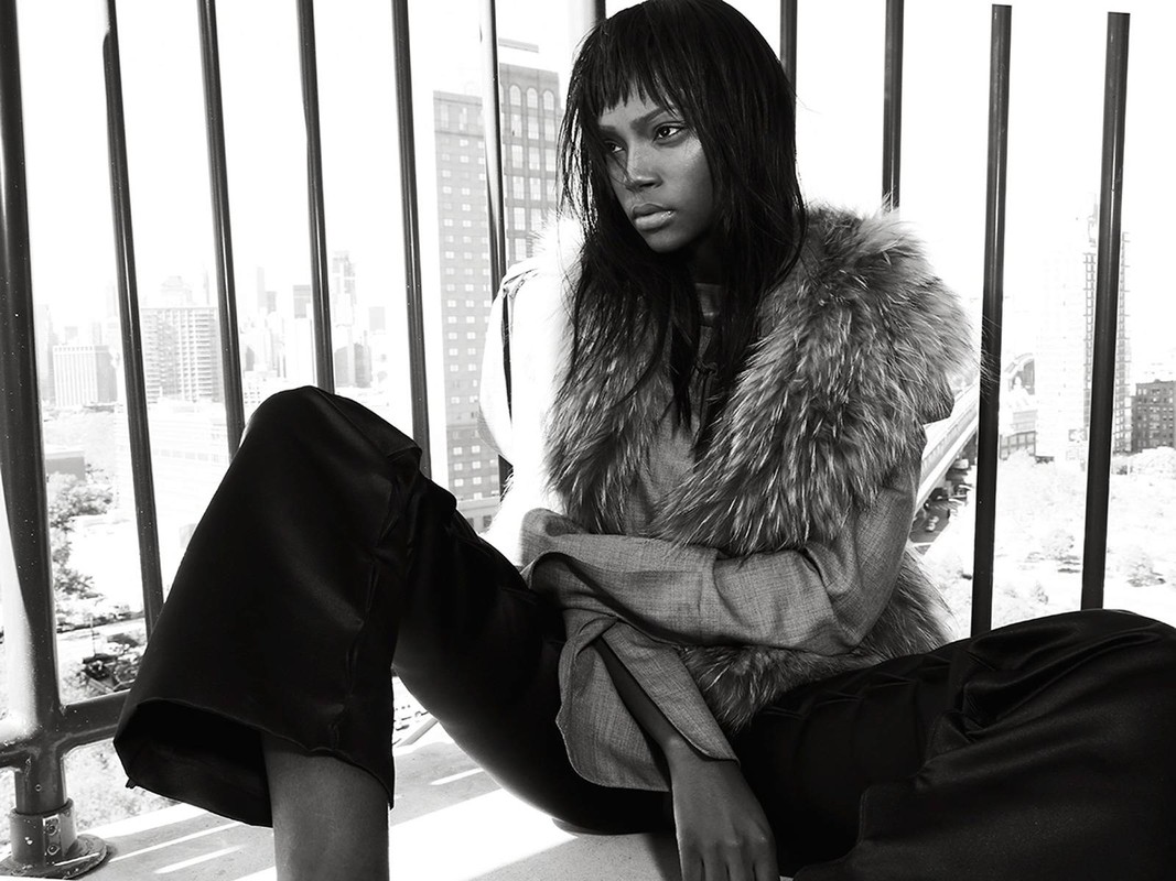 A stylish woman with an urban backdrop, showcasing fashion with a fur collar, wide-leg pants, and a melancholic expression.