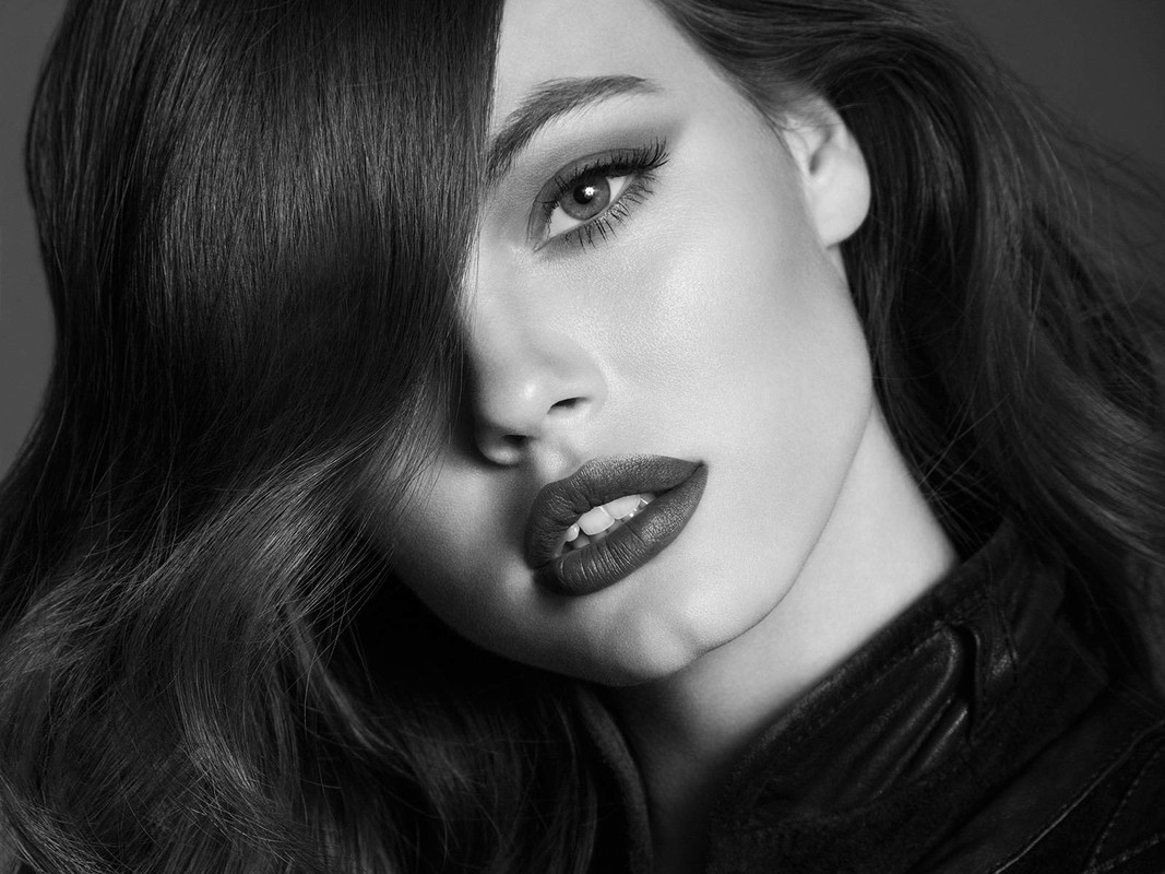 Close-up portrait of a woman with long, wavy black hair partially covering her face, wearing dark lipstick and a leather jacket, in a studio.