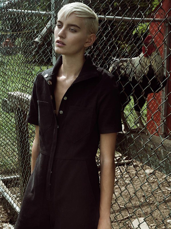 A fair-skinned woman with short, light hair poses in a black jumpsuit, standing outdoors by a chain-link fence with a rooster in the background.