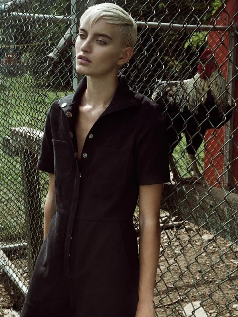 A fair-skinned woman with short, light hair poses in a black jumpsuit, standing outdoors by a chain-link fence with a rooster in the background.