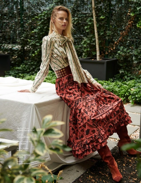 Fashion model in a cream patterned blouse with a ribbon tie and a rust-colored skirt and boots sits outside on a white table.