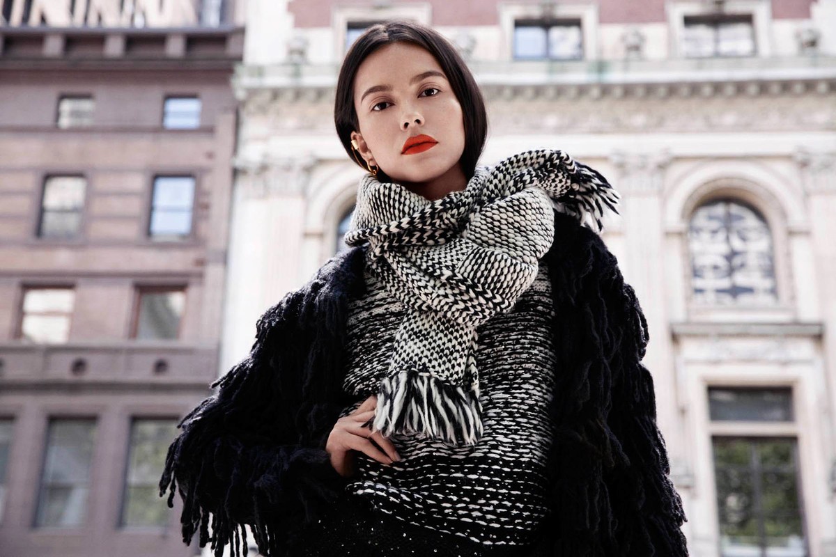 Woman wearing a black and white patterned scarf and fringed jacket outdoors with classic architecture background.