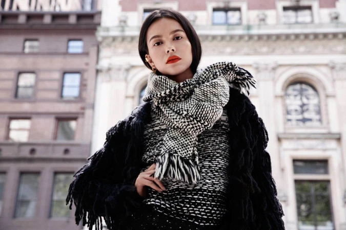 Woman wearing a black and white patterned scarf and fringed jacket outdoors with classic architecture background.