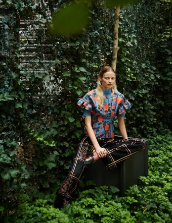 Model with blonde hair wearing a patterned blue top and black trousers seated outdoors among lush green ivy and foliage.