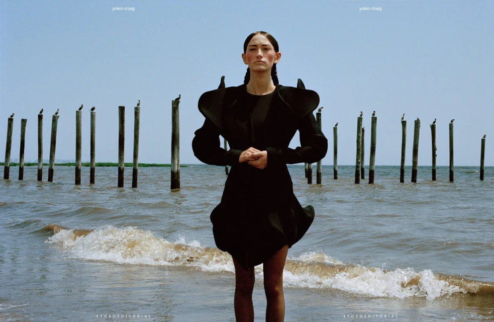 Woman in avant-garde black dress stands on a beach with pilings in the water under a bright sky in a fashion editorial shot.