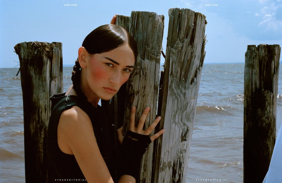 Portrait of a woman with wet slicked-back hair, posing next to weathered wooden posts on a beach with water and sky in the background.