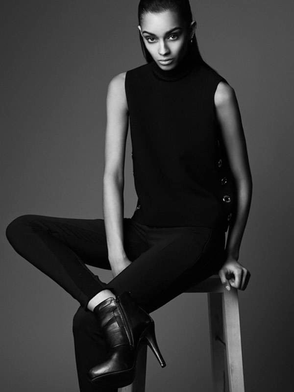 Fashion shot in grayscale featuring a slender woman in a sleeveless top, pants, and heeled boots, posing seated on a wooden stool.