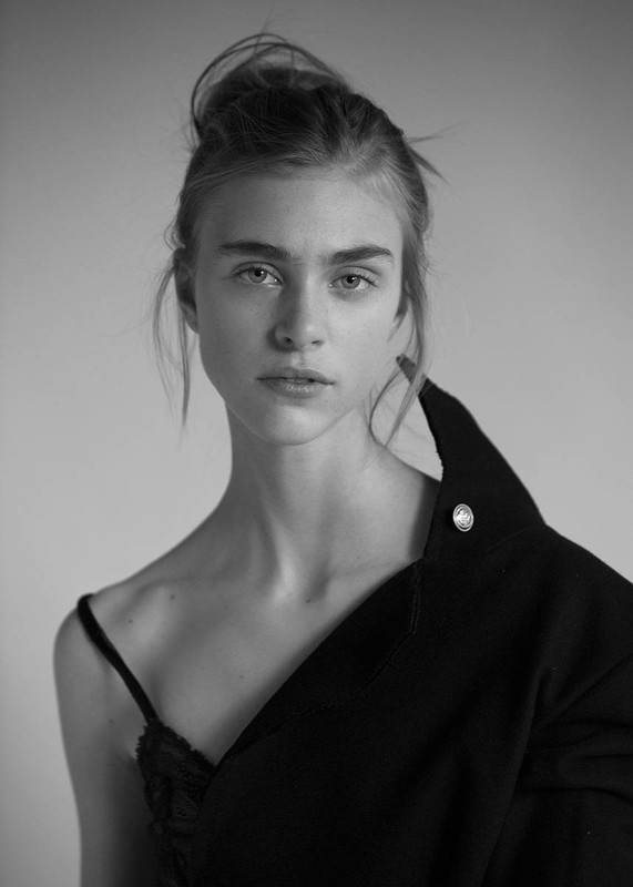 Monochrome portrait of a young woman with hair in a messy bun, wearing a black top and jacket draped over her shoulder.