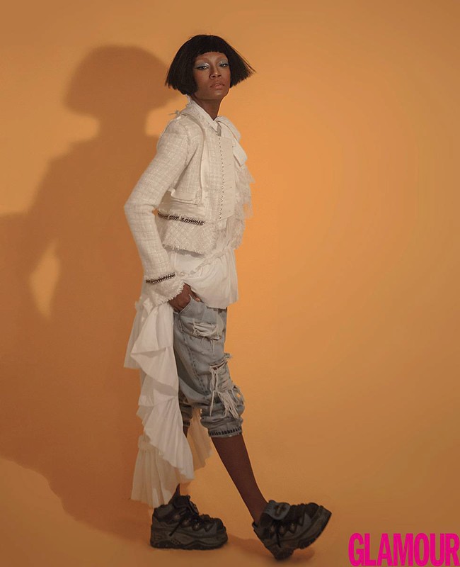 Full-length shot of an adult female model with dark hair posing in a white jacket and platform shoes against a yellow background.