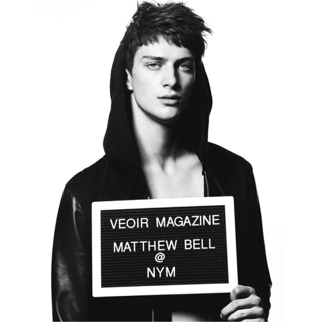 Black and white portrait of a young adult male holding a sign for Veoir Magazine, Matthew Bell, and NYM against a white backdrop.