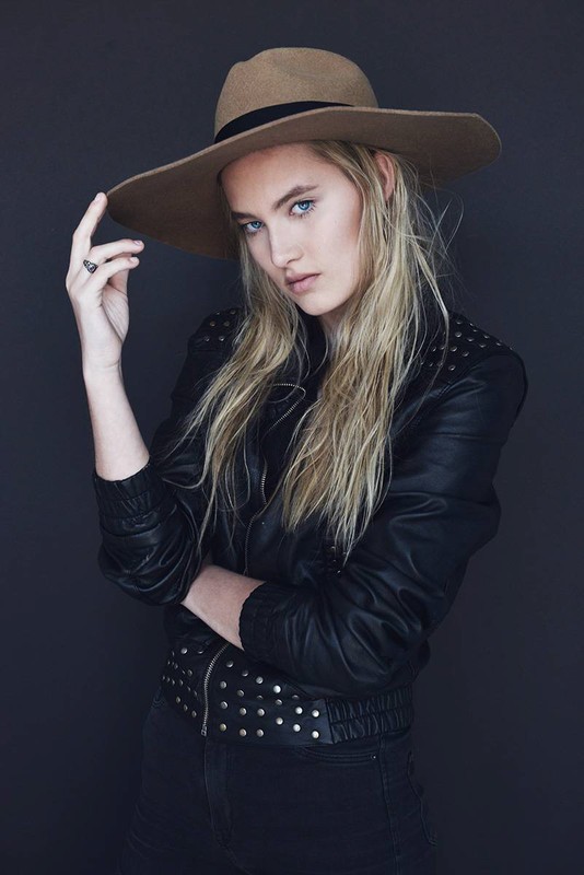Portrait of a blonde woman wearing a hat and black studded jacket against a gray background in a studio setting.