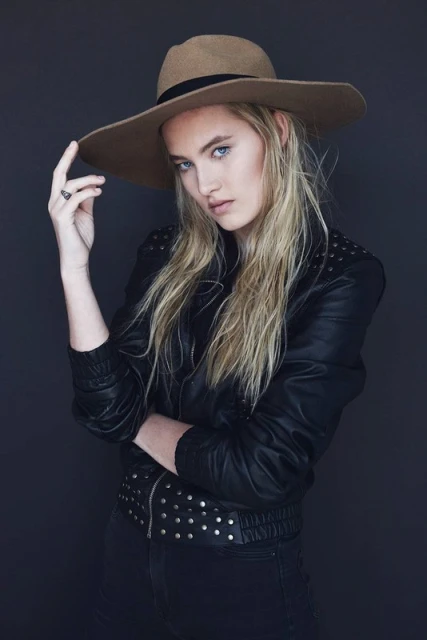 Portrait of a blonde woman wearing a hat and black studded jacket against a gray background in a studio setting.