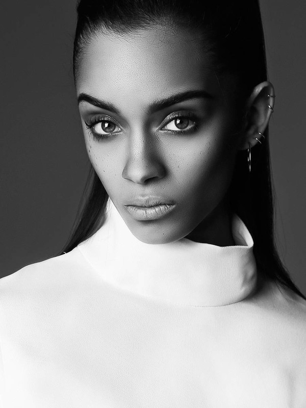 Striking black and white portrait of a woman with dark hair, wearing a white turtleneck sweater against a solid gray background.
