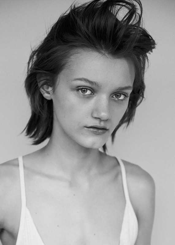 Black and white portrait of a young woman with short, tousled dark hair wearing a light-colored top against a neutral studio backdrop.