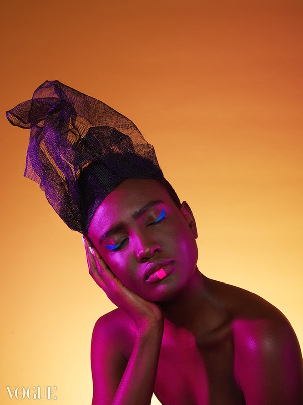 Close-up of an adult with eyes closed, wearing a black head covering and blue eyeliner, skin glowing with pink and orange lighting.