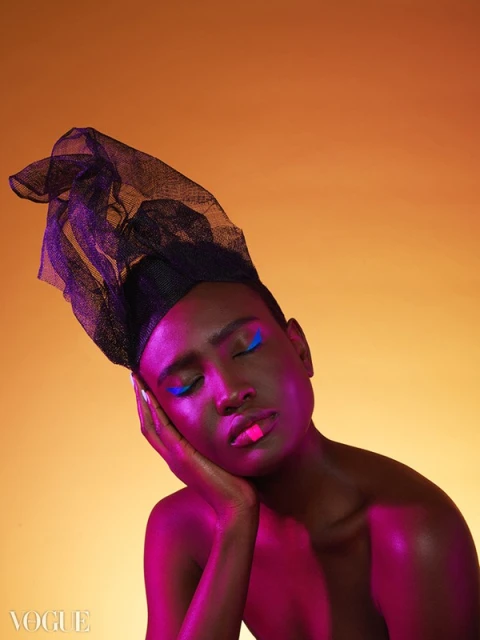 Close-up of an adult with eyes closed, wearing a black head covering and blue eyeliner, skin glowing with pink and orange lighting.