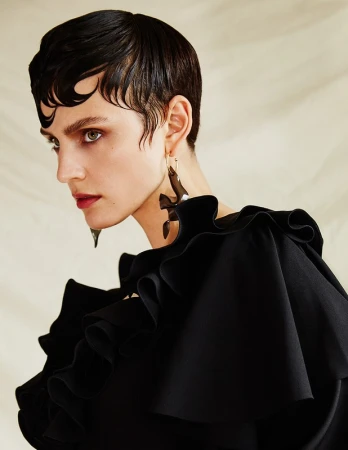 Close-up portrait of a woman with wet-look dark hair and sculptural earrings wearing a ruffled black top against a pale textured background