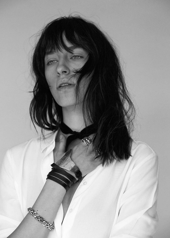 Black and white portrait of a brunette woman in a white shirt, wearing dark bracelets and a ring, hand near her face, neutral expression.