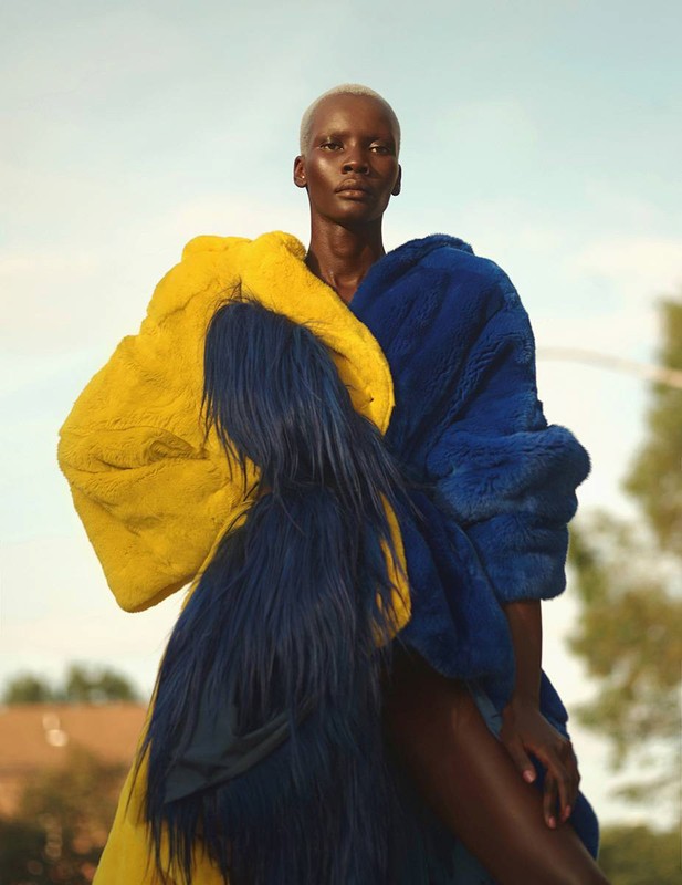 Dark-skinned model wears a large, furry color block coat in yellow and blue outdoors against a pale sky with distant green trees.