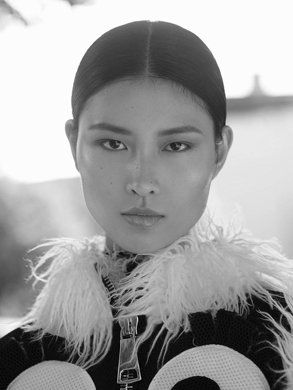 Monochrome portrait of a poised Asian woman with slicked-back hair, wearing a fuzzy-collared jacket and looking directly at the camera.
