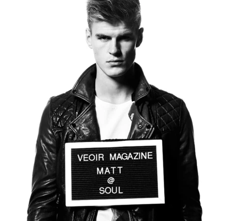 Portrait of a fair-skinned male model with short blond hair, wearing a leather jacket and holding a sign with magazine branding.