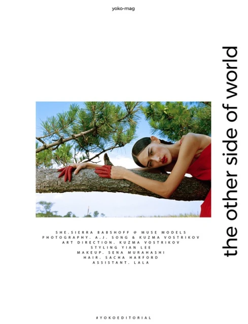An adult woman in red dress and gloves rests on a tree trunk in a bright outdoor fashion editorial, with green pine branches overhead.