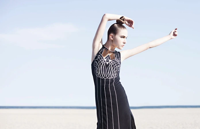 A woman in a black patterned dress with arms raised outdoors against a pale sky and flat sandy ground.