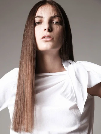 Portrait of a woman with long brown hair wearing a white top against a grey background in a studio setting. Hair and fashion focused.