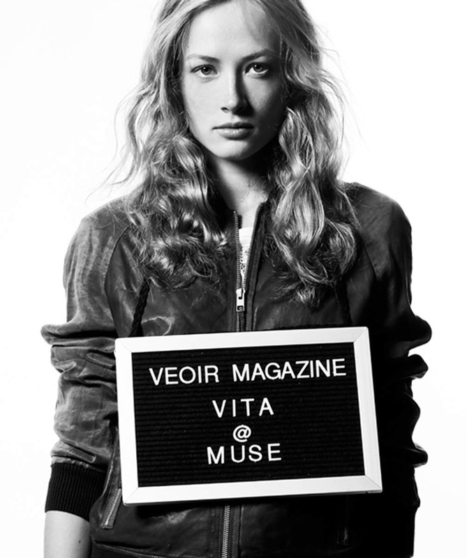 Black and white studio portrait of an adult female model holding a sign with the words 'VEOIR MAGAZINE VITA @ MUSE' on it.