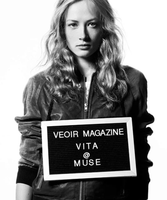 Black and white studio portrait of an adult female model holding a sign with the words 'VEOIR MAGAZINE VITA @ MUSE' on it.