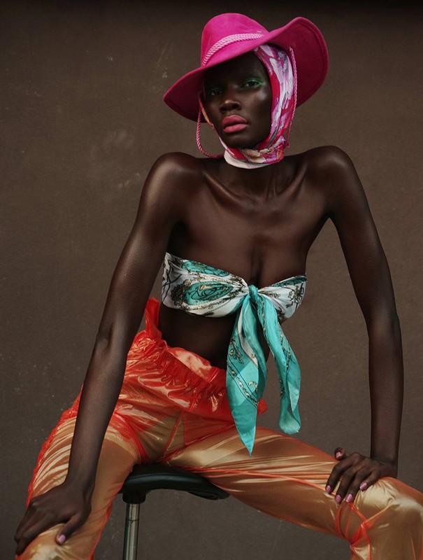 Fashion portrait of a Black woman wearing a pink hat, colorful headscarf, tied top, and orange pants, seated against a brown backdrop.