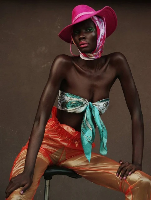 Fashion portrait of a Black woman wearing a pink hat, colorful headscarf, tied top, and orange pants, seated against a brown backdrop.