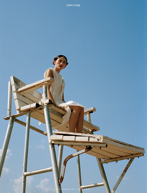 A woman in a white dress sits on a lifeguard chair against a blue sky in an outdoor editorial fashion shot featuring bright, natural lighting.
