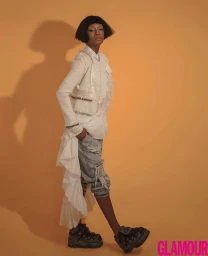 Black woman with short bob hairstyle wearing a white tweed jacket and distressed jeans against an orange backdrop
