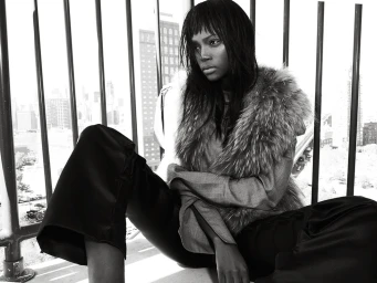 Black and white portrait of a woman with dark hair wearing a fur vest and dark pants seated on a balcony in Los Angeles