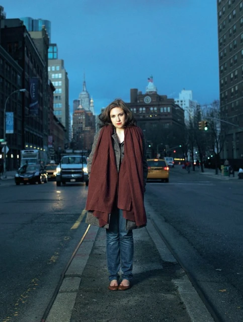 Woman in a maroon scarf stands on a city street in front of buildings and a twilight sky with cars in the background.