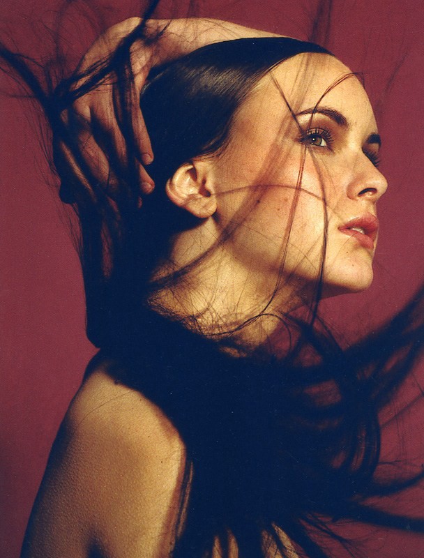 Portrait of an adult woman with dark hair, red lipstick, and light skin, posed against a red backdrop with a hand above her head.
