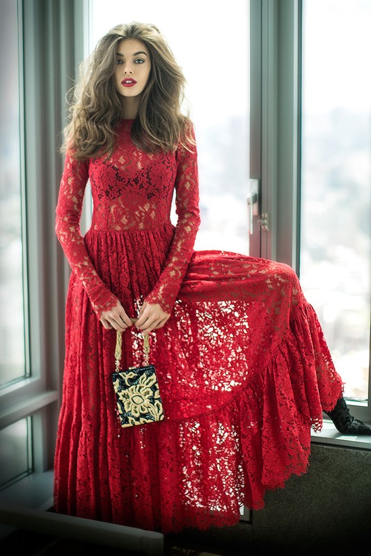 A woman with long brown hair sits on a windowsill wearing a long-sleeved red lace gown and holding a small beaded purse.