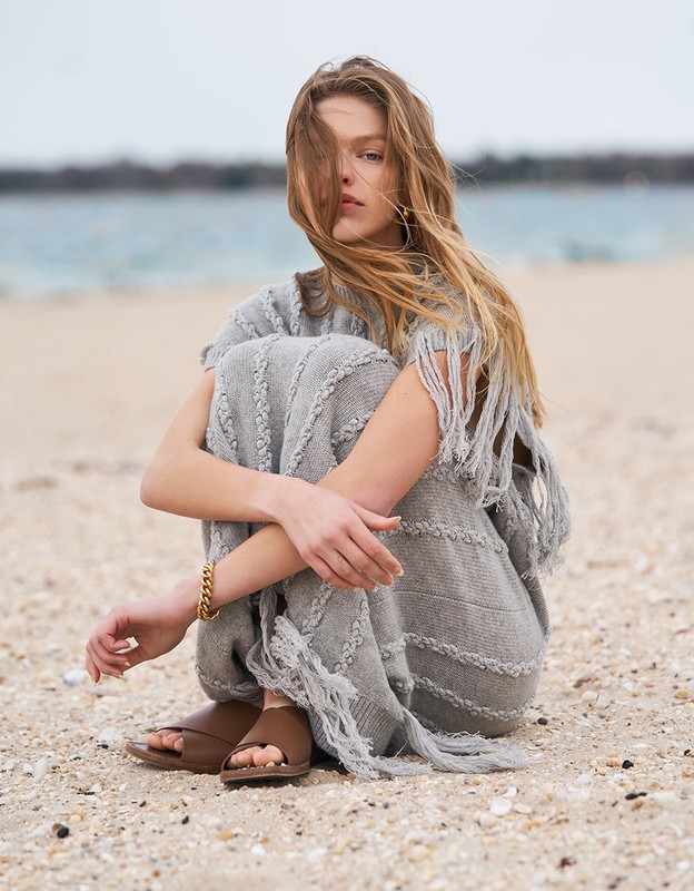 A young woman in a gray, fringed dress sitting on a sandy beach near the ocean on a cloudy day, looking calmly at the camera.