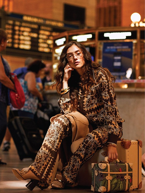 A brunette woman in patterned loungewear poses seated on luggage inside a train station with people and schedule board in the background.